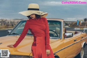 A woman in a red dress and straw hat leaning against a yellow car.