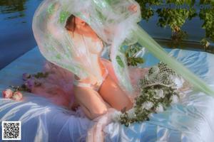 A woman in a pink dress sitting on a blanket by a pond.